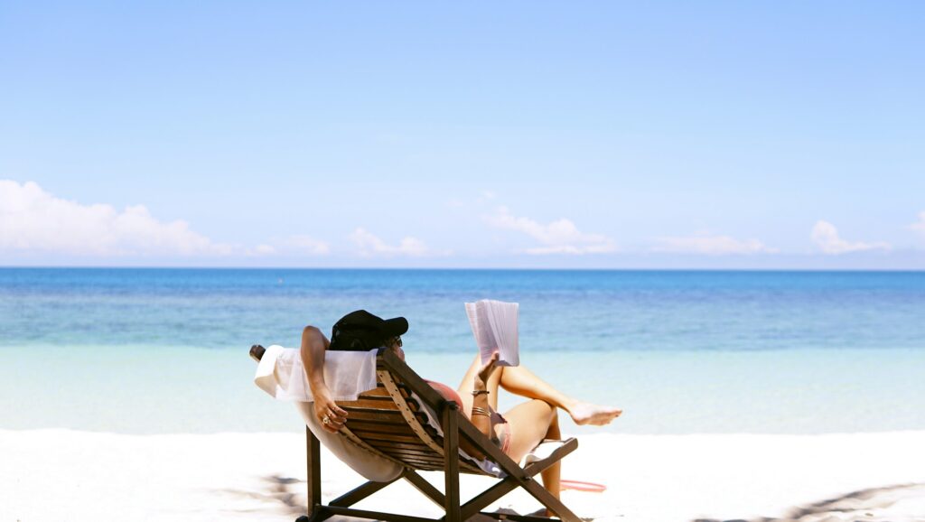 A girl relaxing on a sunbed and reading a book on a sandy beach, with a clear blue sky and turquoise water in the background.